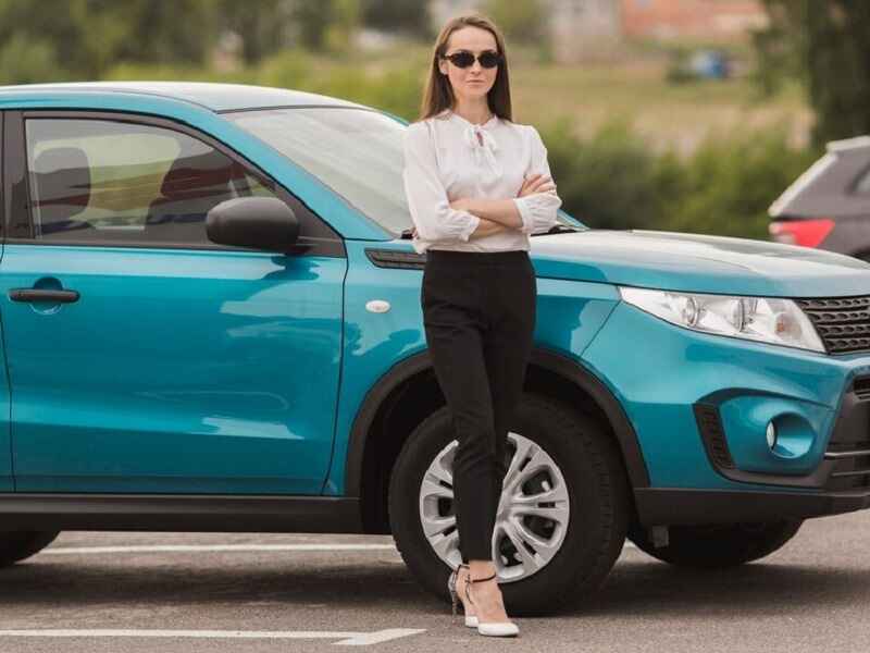 A woman stands next to a blue car with her arms crossed, wearing sunglasses and business clothes. It's in a parking lot with other cars around.