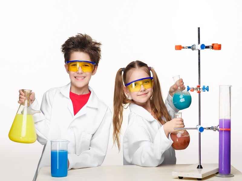 "Two young children wearing lab coats and safety goggles conducting a science experiment. They are smiling while holding flasks filled with colorful liquids, demonstrating curiosity and enthusiasm for science."