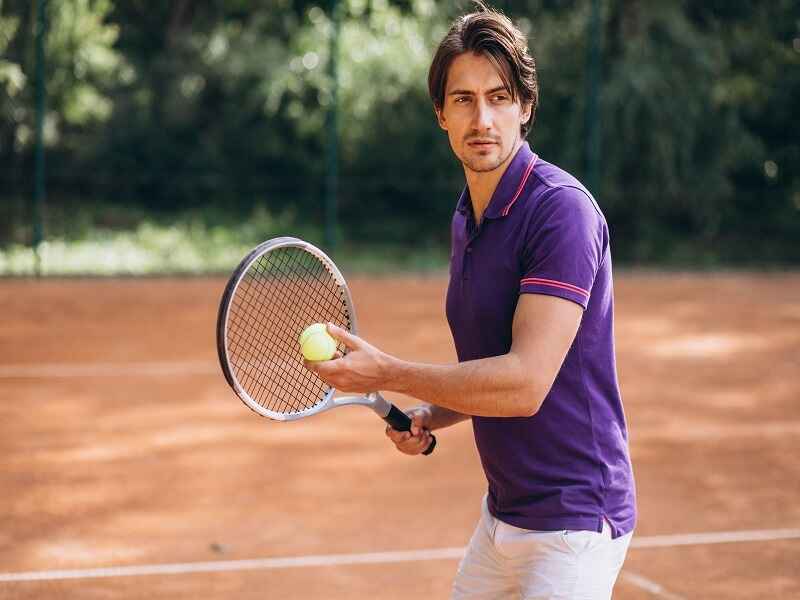  "A man holding a tennis racket and preparing to serve on a clay tennis court, dressed in a purple polo shirt, showcasing focus and readiness."