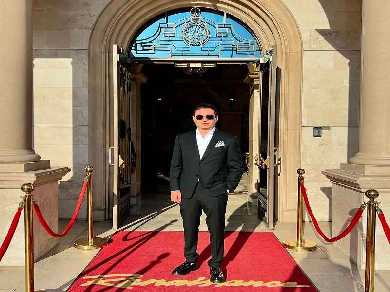 A well-dressed individual standing confidently on a red carpet in front of an elegant building entrance, exuding sophistication and style.