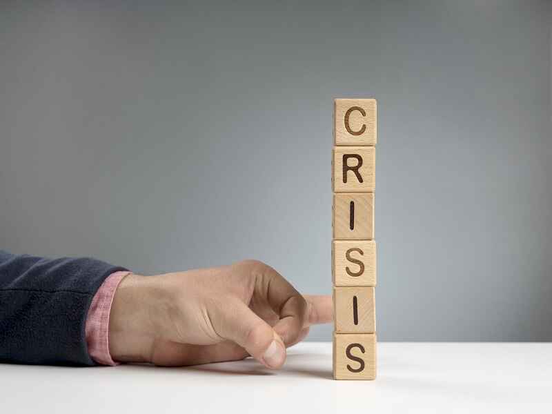 "Hand pushing a stack of wooden blocks spelling 'CRISIS,' symbolizing the delicate balance and risk management involved in preventing business crises or handling critical situations." 