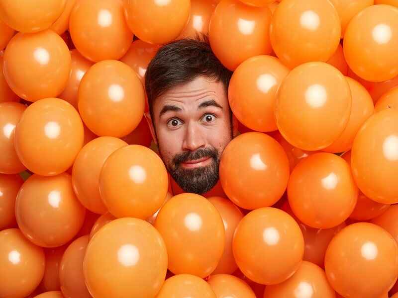 A man with a playful expression is surrounded by a cluster of bright orange balloons, highlighting the vibrancy of the color.