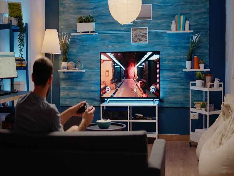 A person sits on a couch in a cozy living room, playing a video game on a big TV. The room has a blue wall, shelves with plants, books, and decor. A lamp gives warm light, and a desk with a computer is on the side. The space feels relaxed and comfy for gaming.