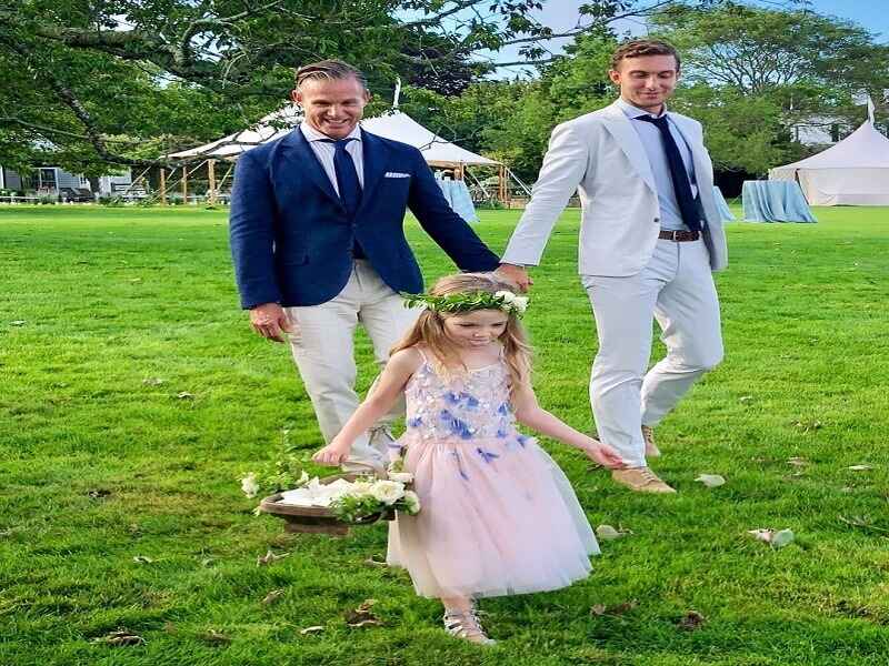 "Two men in formal attire smiling and holding hands, walking on a grassy area alongside a young girl dressed in a floral crown and light pink dress, carrying a basket of flowers, with a tent visible in the background."