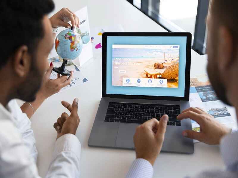 "Two men planning a trip using a laptop with a travel website on the screen, emphasizing the importance of researching and booking travel accommodations in advance for a smooth journey."