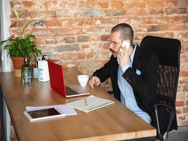 "Man engaged in a phone call while working at his desk, focusing intently on his laptop and notes, representing the concept of active listening and mindful observation in a professional setting."