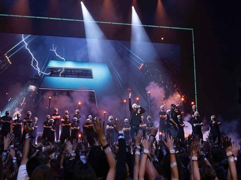 "Eminem performing on stage in front of a large crowd, with a dramatic background featuring lightning and a large letter 'E,' symbolizing his iconic presence and energy in the music industry."